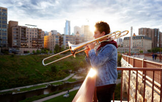 Portrait of a high school senior play his trombone on a bridge with an urban skyline in background