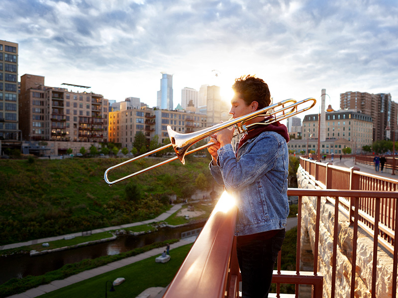 Portrait of a high school senior play his trombone on a bridge with an urban skyline in background